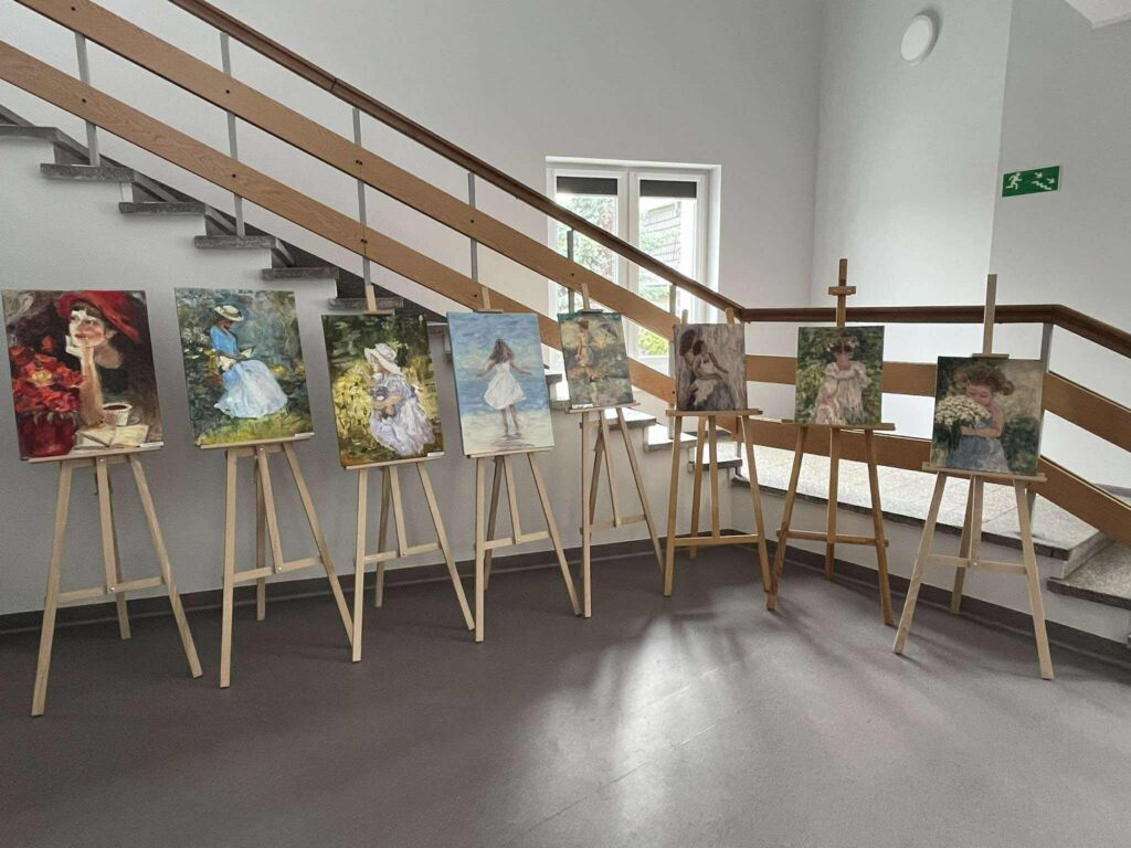 W holu biblioteki stoją drewniane sztalugi z obrazami przedstawiającymi dzieci.