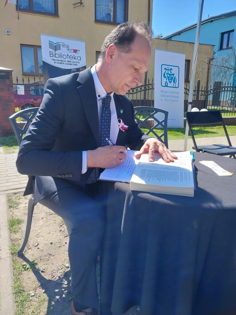 W plenerze przy okrągłym stoliku nakrytym czarnym obrusem mieszkańcy przepisują do otwartej księgi wersy Pana Tadeusza.