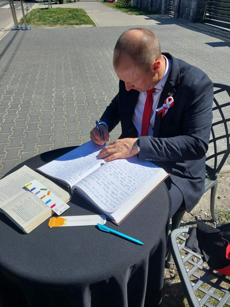 W plenerze przy okrągłym stoliku nakrytym czarnym obrusem mieszkańcy przepisują do otwartej księgi wersy Pana Tadeusza.