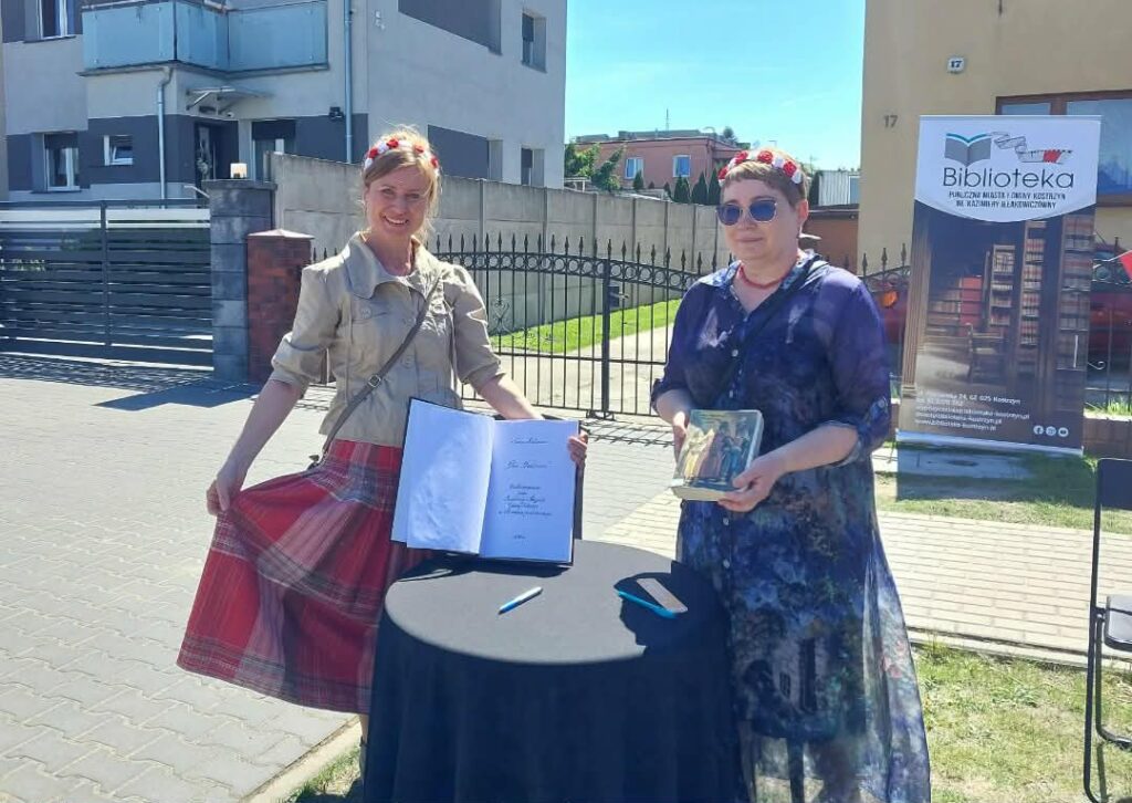 Dwie kobiety w biało-czerwonych wiankach na głowie stoją na trawniku przy okrągłym stoliku.