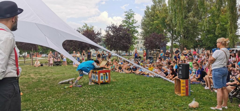 Publiczność w parku ogląda spektakl na pierwszym planie mim nachyla się nad kolorową skrzynią