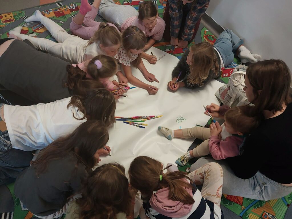 Na dywanie wokół arkusza papieru leżą dzieci z pisakami w dłoniach i rysują.