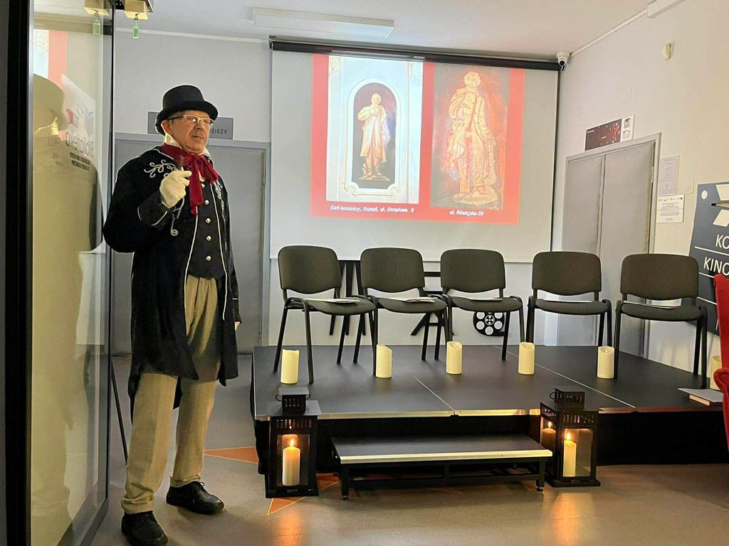 Na pierwszym planie po lewej mężczyzna w czarnym cylindrze w ubraniu stylizowanym na odświętny strój z epoki romantyzmu w tle krzesła na scenie oraz ekran z grafiką prezentacji