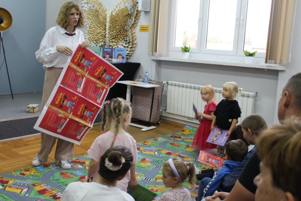 w CZYTELNI NA KOLOROWYM DYWANIE SIEDZĄ DZIECI. kOBIETA W BIAŁEJ BLUZCE TRZYMA CZERWONY ARKUSZ PAPIERU.