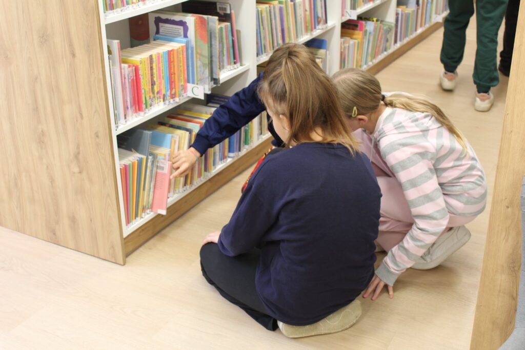 Uczennice szukają książki na dolnej półce regału bibliotecznego.